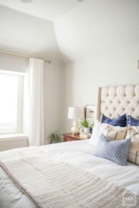 Welcome to our Spring Home Tour: See how we transformed our home with just a few simple changes, a modern farmhouse master bedroom with white curtains, white bedding, dark nightstand with white gourd lamp and greenery a mix of throw pillows and a neutral quilt create calming enviroment in this space.