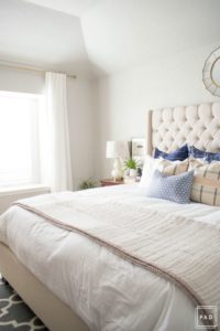 Welcome to our Spring Home Tour: See how we transformed our home with just a few simple changes, a modern farmhouse master bedroom with white curtains, white bedding, dark nightstand with white gourd lamp and greenery a mix of throw pillows and a neutral quilt create calming enviroment in this space.