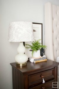 Welcome to our Spring Home Tour: See how we transformed our home with just a few simple changes, a modern farmhouse master bedroom with white curtains, white bedding, dark nightstand with white gourd lamp and greenery a mix of throw pillows and a neutral quilt create calming enviroment in this space.